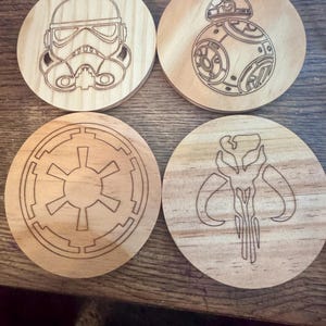 Puede incluir: Juego de cuatro posavasos de madera con diseños grabados. Incluye un casco de Stormtrooper, el droide BB-8, el símbolo del Imperio Galáctico y una calavera mandaloriana. Los posavasos son redondos y de madera clara.