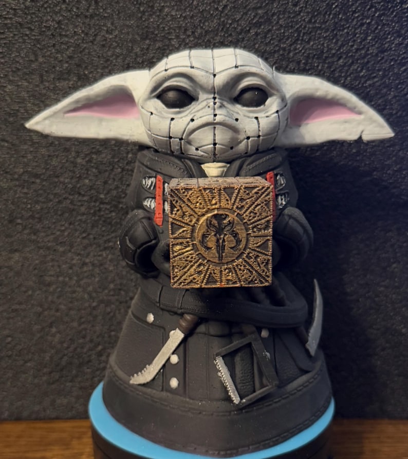 May include: A small, white and grey figurine of a character from the Star Wars franchise, holding a golden box with a Mandalorian symbol. The figurine is wearing a black jacket with a red and white design.