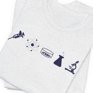 May include: A light gray t-shirt featuring a series of navy blue science-themed graphics, including a syringe, atom, petri dish, flask, and microscope. The shirt is folded, showing the front design.