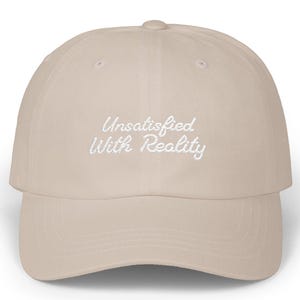 Puede incluir: Gorra de béisbol beige con visera curva. La gorra presenta el texto bordado "Unsatisfied With Reality" en blanco y cursiva. El sombrero está hecho de un material suave y tiene un estilo clásico e informal.
