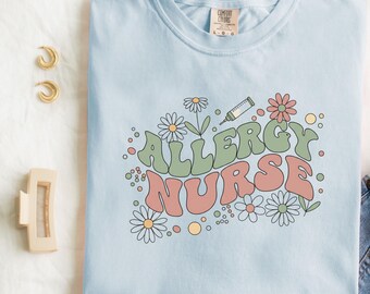 以下が含まれることがあります： レトロなフォントで「ALLERGY NURSE」と書かれた水色のTシャツ。デイジーと花のグラフィック、注射器のイラストが描かれています。Tシャツは白い表面に折りたたまれ、ジーンズ、イヤリング、ヘアクリップの隣に置かれています。