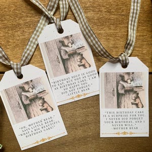 May include: Three gift tags with illustrations of bears and text. Each tag has a different quote about birthdays. The tags are tied with a brown and white checkered ribbon. The background is a wooden surface.