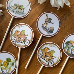 May include: A selection of round cupcake toppers with illustrations of frogs in different scenarios. Each topper has a brown border and is attached to a white stick. Ideal for a themed celebration.
