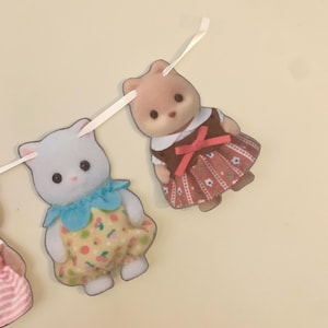 Pode incluir: Uma guirlanda com duas figuras de animais de brinquedo. Uma &eacute; um gato branco usando um vestido amarelo com um padr&atilde;o floral e uma gola azul. A outra &eacute; um urso marrom usando um vestido marrom com um la&ccedil;o vermelho.