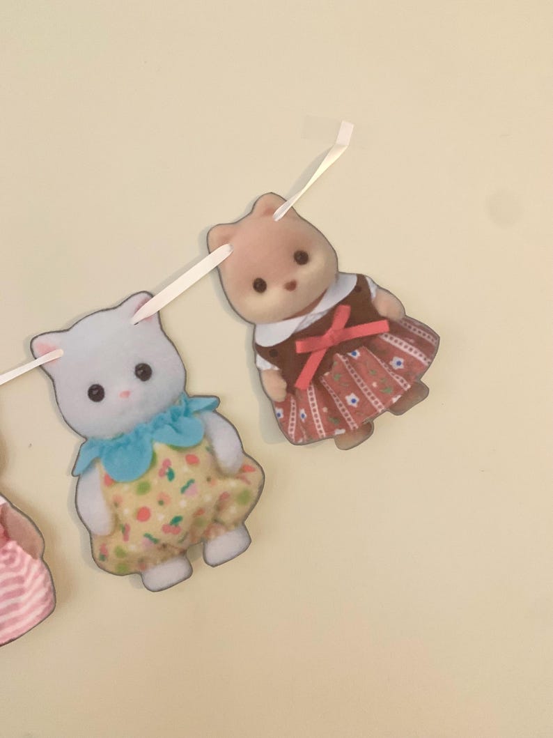 Pode incluir: Uma guirlanda com duas figuras de animais de brinquedo. Uma &eacute; um gato branco usando um vestido amarelo com um padr&atilde;o floral e uma gola azul. A outra &eacute; um urso marrom usando um vestido marrom com um la&ccedil;o vermelho.