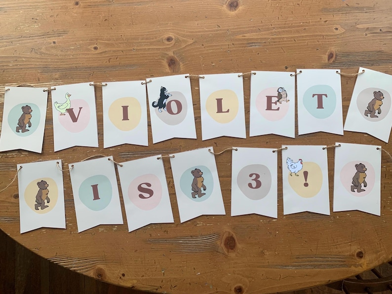 May include: A banner spelling "VIOLET IS 3" with individual pennants. Each pennant has a letter or number, and a corresponding animal illustration. The banner is strung on twine and displayed on a wooden table.