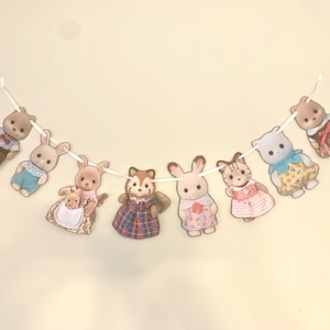 Pode incluir: Uma guirlanda decorativa com nove figuras de animais, incluindo coelhos e um esquilo, enfiadas numa fita branca. As figuras usam roupas variadas, com cores como azul, rosa e castanho. Ideal para um quarto infantil.