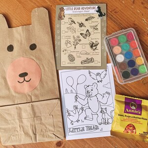 May include: A brown paper bear-shaped bag, a scavenger hunt card, a watercolor paint set, a coloring page, and a snack bag are arranged on a wooden surface. The scavenger hunt card is titled "Little Bear Adventure". The snack bag is labeled "Annie's Bees, Bugs & Butterflies".