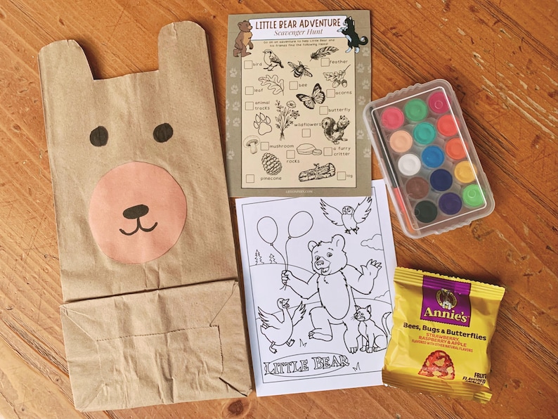 May include: A brown paper bear-shaped bag, a scavenger hunt card, a watercolor paint set, a coloring page, and a snack bag are arranged on a wooden surface. The scavenger hunt card is titled "Little Bear Adventure". The snack bag is labeled "Annie's Bees, Bugs & Butterflies".