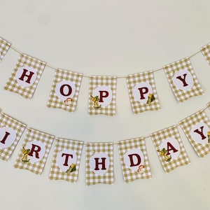 May include: A "Happy Birthday" banner with alternating tan and white checkered flags. Each flag displays a letter in burgundy, with illustrations of a bear riding a bicycle and other woodland creatures. The banner is strung on a thin, natural-colored twine.