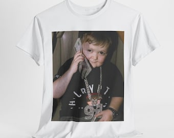 Op de afbeelding: Een wit T-shirt met een foto van een jonge jongen die een grote gouden ketting draagt en een stapel geld aan zijn oor houdt. De jongen draagt een zwart shirt met de tekst "H.I. CRYPT" en "94" erop gedrukt.