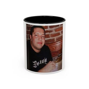 以下が含まれることがあります： 黒い縁取りのある白いセラミック製のマグカップ。男性が「デトロイト」と書かれた黒いシャツを着て、レストランに座って料理の入った皿を持っている写真がプリントされています。