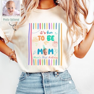 May include: An ivory short-sleeve t-shirt featuring a colorful graphic with vertical stripes and the text 'it's fun TO BE MOM' and 'Oliver's First Birthday'. The design includes illustrations of a cake, gift, and balloons, with a baby photo.