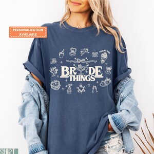 May include: A navy blue t-shirt with the words "BRIDE THINGS" in a white, stylized font, along with various white illustrations. The shirt has rolled-up sleeves and is paired with a denim jacket. The text "PERSONALIZATION AVAILABLE" is in an orange box.