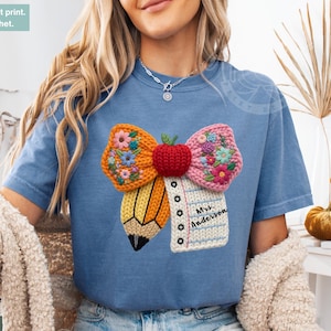 May include: Blue jean color t-shirt featuring a colorful patch-effect design of a pencil, apple, and bow with floral accents. The design includes the text "Mrs. Anderson" on a notepad. The shirt has a crew neck and short sleeves.