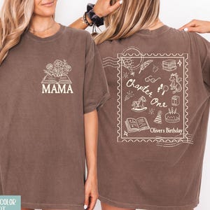 May include: Espresso-colored t-shirt with a front graphic of an open book with flowers and the word "MAMA" in white. The back features a white illustration with the words "Chapter One" and "Oliver's Birthday".