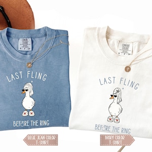 May include: Two t-shirts, one blue and one ivory, featuring a cartoon duck wearing sunglasses and a towel, with the text "LAST FLING BEFORE THE RING." Each shirt has a gold necklace. The blue shirt is labeled "BLUE JEAN COLOR T-SHIRT" and the ivory shirt is labeled "IVORY COLOR T-SHIRT."