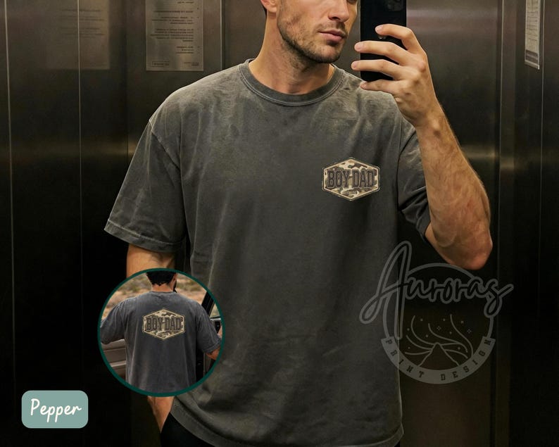 May include: Pepper-colored, oversized t-shirt with a faded, vintage look. The shirt features a camouflage-patterned, hexagonal patch that reads "BOY DAD". The shirt is a casual wear item.