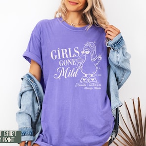 May include: A violet t-shirt with ivory print. The shirt displays the text "GIRLS GONE Mild" and a cartoon duck wearing sunglasses and holding a drink. The text "Amanda's bachelorette + Chicago, Illinois" is also printed on the shirt.