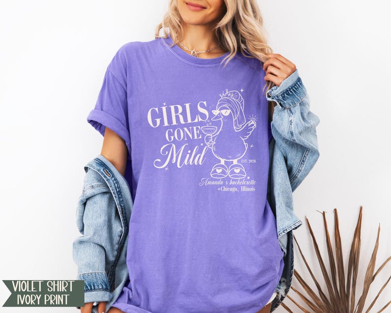 May include: A violet t-shirt with ivory print. The shirt displays the text "GIRLS GONE Mild" and a cartoon duck wearing sunglasses and holding a drink. The text "Amanda's bachelorette + Chicago, Illinois" is also printed on the shirt.