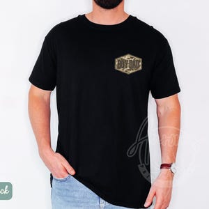 May include: Black t-shirt featuring a small, camouflage-patterned patch with the text "BOY DAD" in gold. The shirt has a crew neck and short sleeves. The model is wearing blue jeans and a wristwatch.