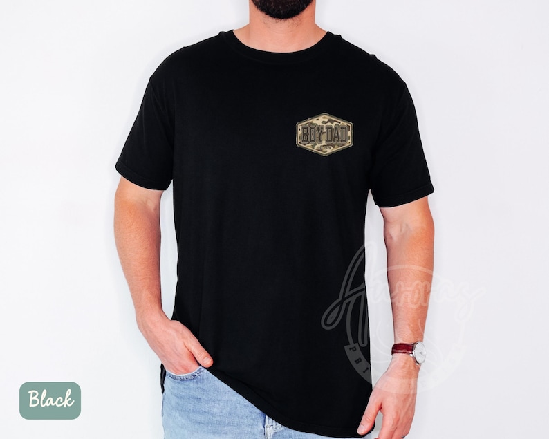 May include: Black t-shirt featuring a small, camouflage-patterned patch with the text "BOY DAD" in gold. The shirt has a crew neck and short sleeves. The model is wearing blue jeans and a wristwatch.