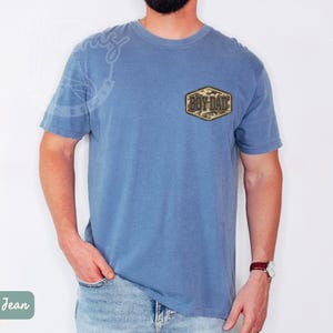 May include: Blue short-sleeved t-shirt with a camouflage patch that reads "Boy Dad." The shirt is a solid blue colour and has a relaxed fit. The model is wearing blue jeans and a watch.