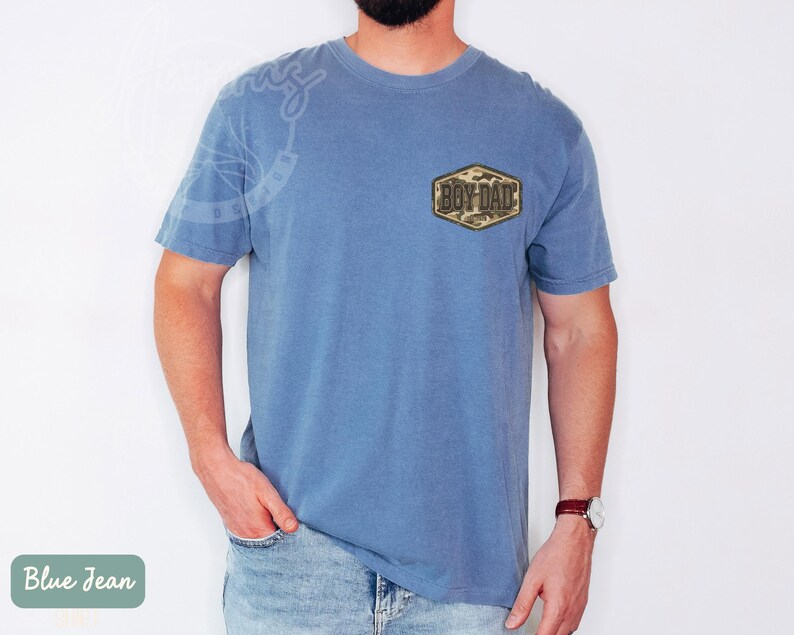 May include: Blue short-sleeved t-shirt with a camouflage patch that reads "Boy Dad." The shirt is a solid blue colour and has a relaxed fit. The model is wearing blue jeans and a watch.
