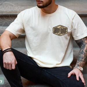 May include: Ivory t-shirt featuring a small camouflage patch with the text "BOY DAD". The short-sleeved shirt has a crew neck. The model is wearing black jeans and a black beaded bracelet.