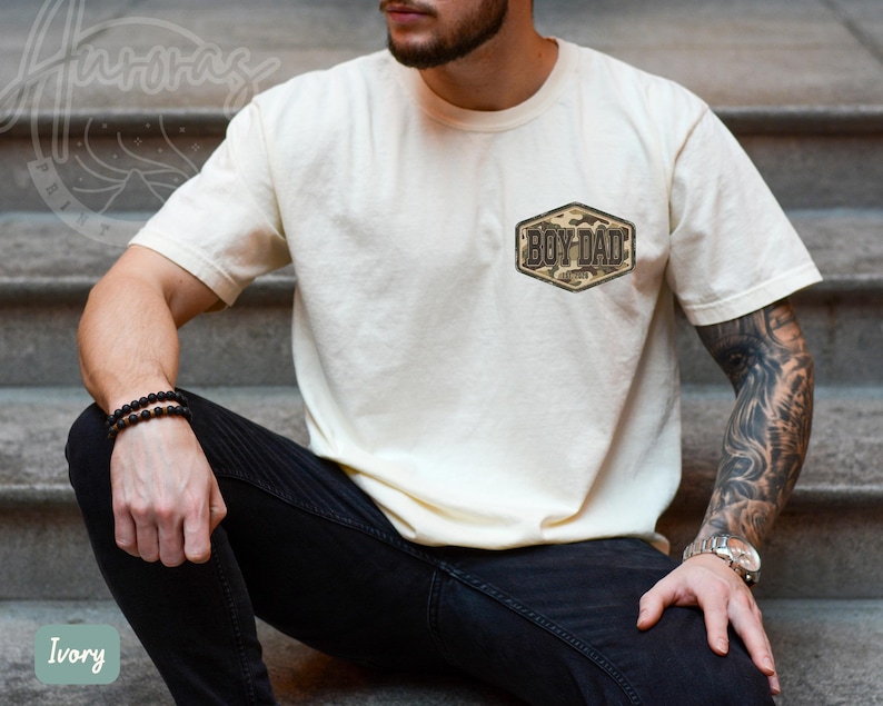 May include: Ivory t-shirt featuring a small camouflage patch with the text "BOY DAD". The short-sleeved shirt has a crew neck. The model is wearing black jeans and a black beaded bracelet.
