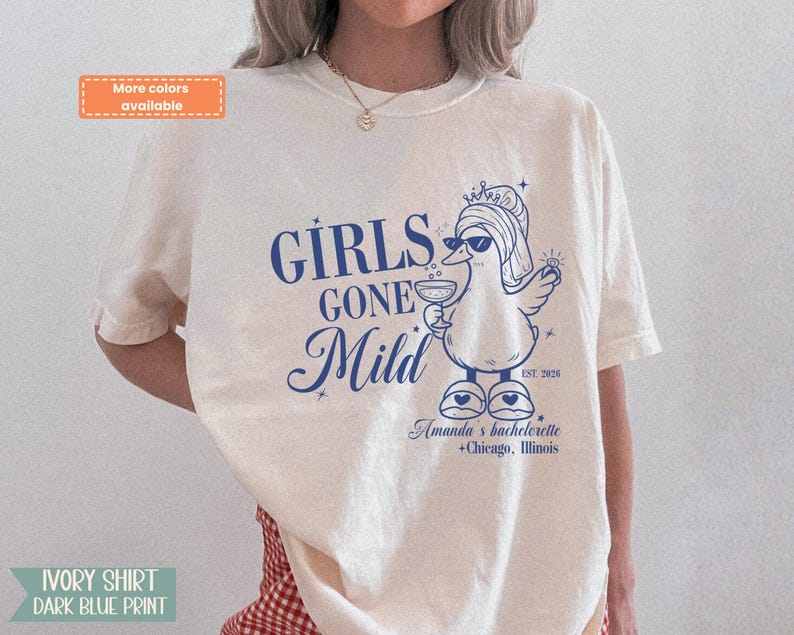May include: Cream-coloured t-shirt with a dark blue graphic. The graphic says "GIRLS GONE Mild" and features a cartoon duck wearing sunglasses and a crown, holding a drink. The text "Amanda's bachelorette + Chicago, Illinois" is below.