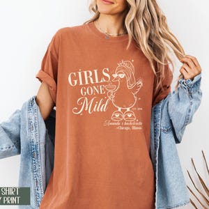 May include: A terracotta-coloured t-shirt with the text "GIRLS GONE Mild" and a cartoon duck wearing a crown and holding a cocktail. The shirt also includes the text "Amanda's bachelorette + Chicago, Illinois".