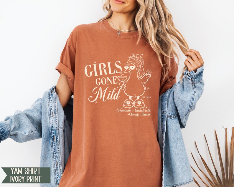 May include: A terracotta-coloured t-shirt with the text "GIRLS GONE Mild" and a cartoon duck wearing a crown and holding a cocktail. The shirt also includes the text "Amanda's bachelorette + Chicago, Illinois".