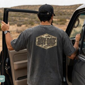 May include: A charcoal grey t-shirt featuring the text "BOY DAD" in a camouflage design. The short-sleeved shirt has a vintage appearance. The person is standing next to a black car.
