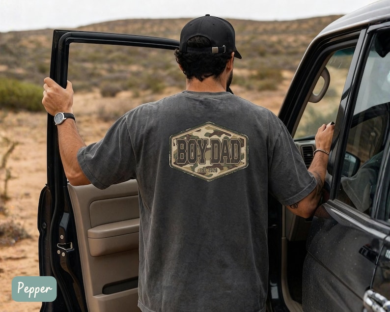 May include: A charcoal grey t-shirt featuring the text "BOY DAD" in a camouflage design. The short-sleeved shirt has a vintage appearance. The person is standing next to a black car.