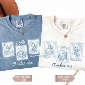 May include: Two t-shirts, one blue jean and one ivory, each with a unique design. The design features a cake, books, and a dragon within postage stamp frames, with the text "Chapter one" below. Both shirts have a gold necklace.