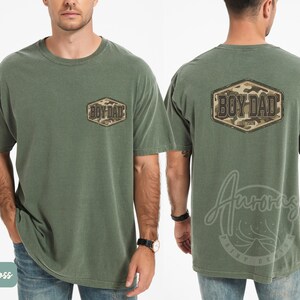 May include: Moss green t-shirt featuring "BOY DAD" text in a camouflage design on the front and back. The shirt has a crew neck and short sleeves. The shirt is made of a soft, comfortable material.