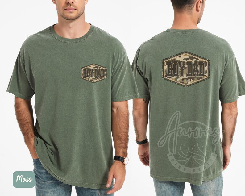 May include: Moss green t-shirt featuring "BOY DAD" text in a camouflage design on the front and back. The shirt has a crew neck and short sleeves. The shirt is made of a soft, comfortable material.