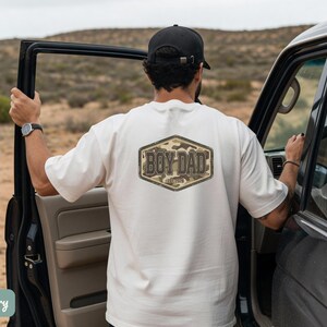 May include: Ivory t-shirt featuring a "BOY DAD" design on the back, set within a hexagonal frame with a camouflage pattern. The person is wearing a black cap and watch, standing beside a black vehicle.