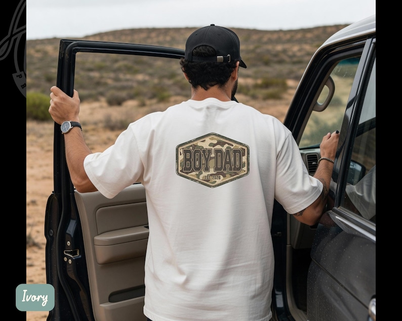 May include: Ivory t-shirt featuring a "BOY DAD" design on the back, set within a hexagonal frame with a camouflage pattern. The person is wearing a black cap and watch, standing beside a black vehicle.
