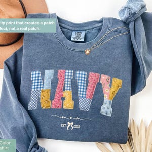 May include: Denim blue sweatshirt with the word "NAVY" in colorful patchwork letters. The sweatshirt has a crew neck and long sleeves. The text "mom EST. 2026" is printed below the word "NAVY". A gold necklace is draped on the sweatshirt.