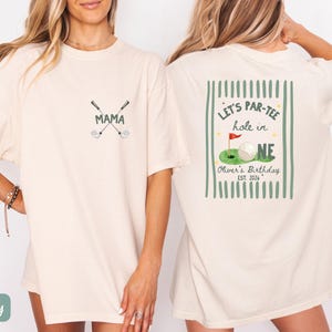 May include: An ivory short-sleeve t-shirt featuring a small graphic on the front with two crossed golf clubs and the word MAMA. The back displays a large graphic with green vertical stripes, a golf ball on a green, and the text LET'S PAR-TEE hole in ONE, Oliver's Birthday, EST. 2026.