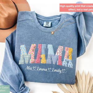 以下が含まれることがあります： パッチワークデザインの「MAMA」という文字が入った、ブルーデニムカラーのスウェットシャツ。文字は花柄とギンガムチェック柄で装飾されています。「MAMA」の下には、「Alex Emma Emily」という名前とハートのシンボルがあります。