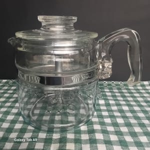May include: A clear glass percolator coffee pot featuring a transparent lid, a sturdy glass handle, and a central metal component with the word STAINLESS embossed on the side. The vessel has a cylindrical body and a wide, flat base design.