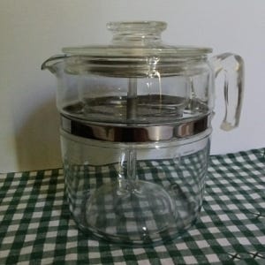 May include: A clear glass coffee percolator with a glass lid and handle. The percolator has a silver metal band around the middle and a glass tube in the centre. The percolator is on a green and white checkered tablecloth.