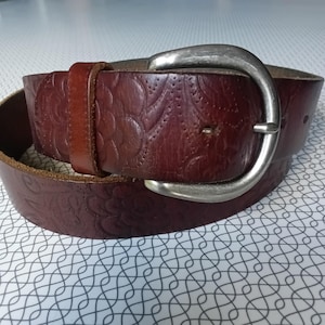 May include: A brown leather belt with a floral embossed pattern. The belt features a silver-coloured buckle and a matching leather loop. The belt is approximately 3.8 cm wide.