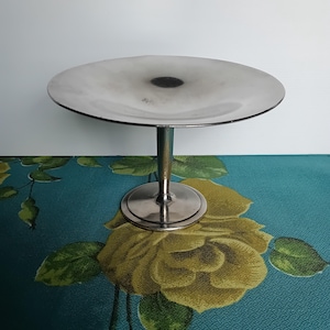 May include: A silver-toned metal pedestal stand featuring a wide, shallow, circular top plate supported by a slender central column and a stable, flat, round base. The surface of the metal has a polished finish with subtle dark shading.