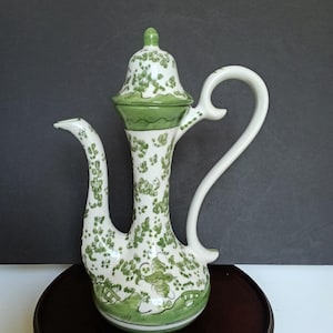 May include: A white ceramic pitcher with a curved spout and handle, adorned with green floral patterns. It features a green band at the base and lid, and a small green knob on the lid. The pitcher is displayed on a dark wooden surface.
