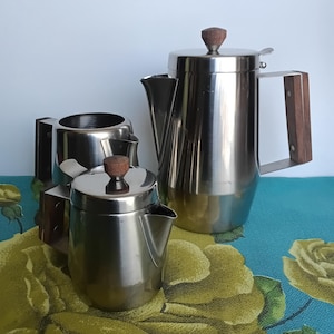 May include: A stainless steel coffee service set consisting of a tall coffee pot, a smaller creamer, and an open sugar bowl. Each piece features a polished metallic finish and dark brown wooden handles, with matching wooden knobs on the lids.