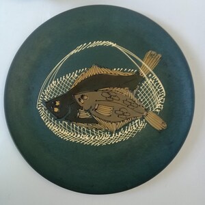 May include: A round, dark teal ceramic plate featuring a hand-painted design of two fish within a white net. The fish are in shades of brown and black, with detailed fins and scales. The plate has a matte finish.